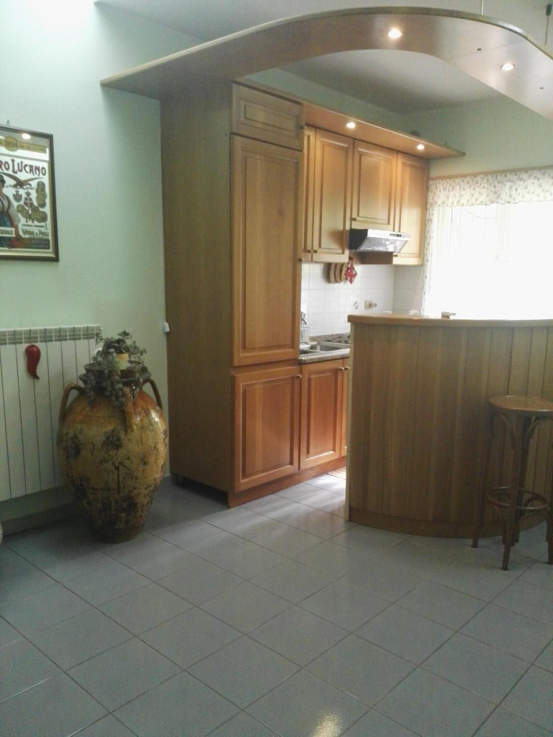 Cocina con alacenas de madera, un jarrón grande y un taburete pequeño.