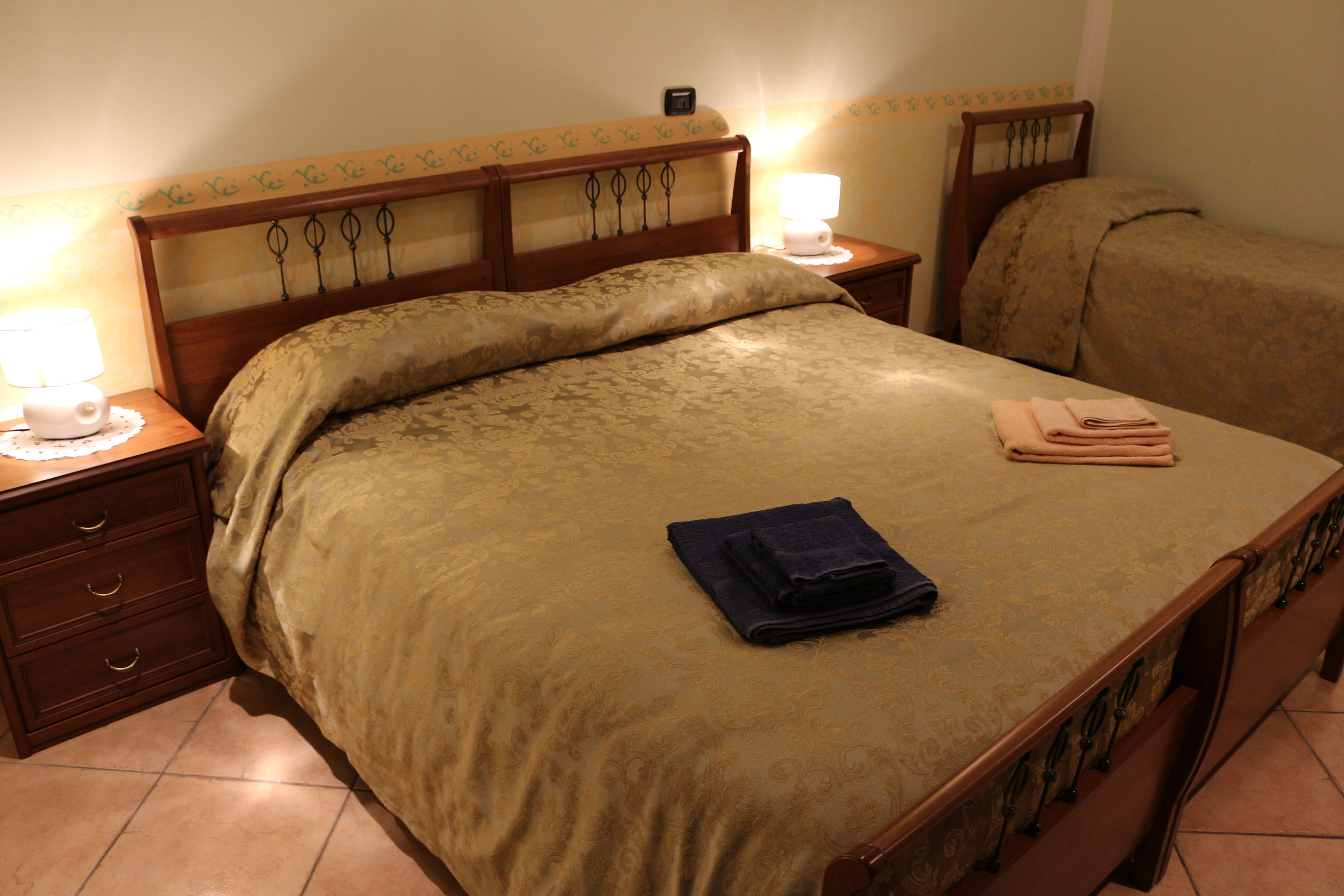 Een net opgemaakt bed met een houten hoofdbord, twee nachtkastjes met lampen, en een opgevouwen handdoek op het bed.