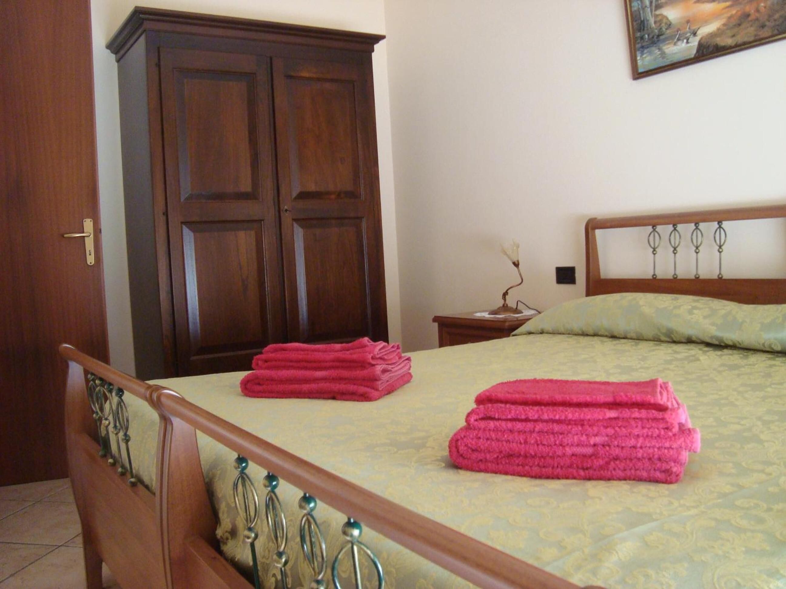 A bedroom with a wooden bed frame, a green patterned bedspread, and two pink towels on the bed. There is a wooden wardrobe and a small bedside table with a lamp.