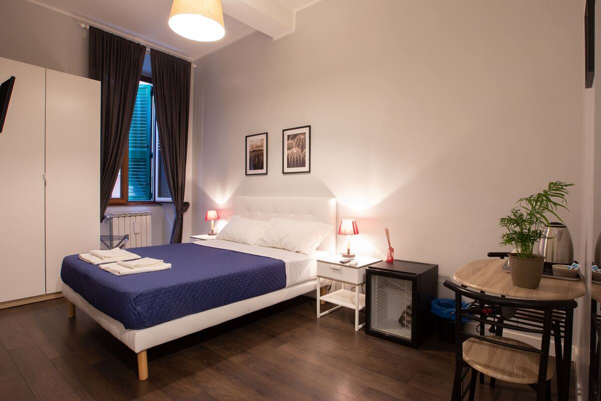 Una habitación de hotel moderna con cama, mesita de noche, lámpara y una pequeña mesa con una planta.