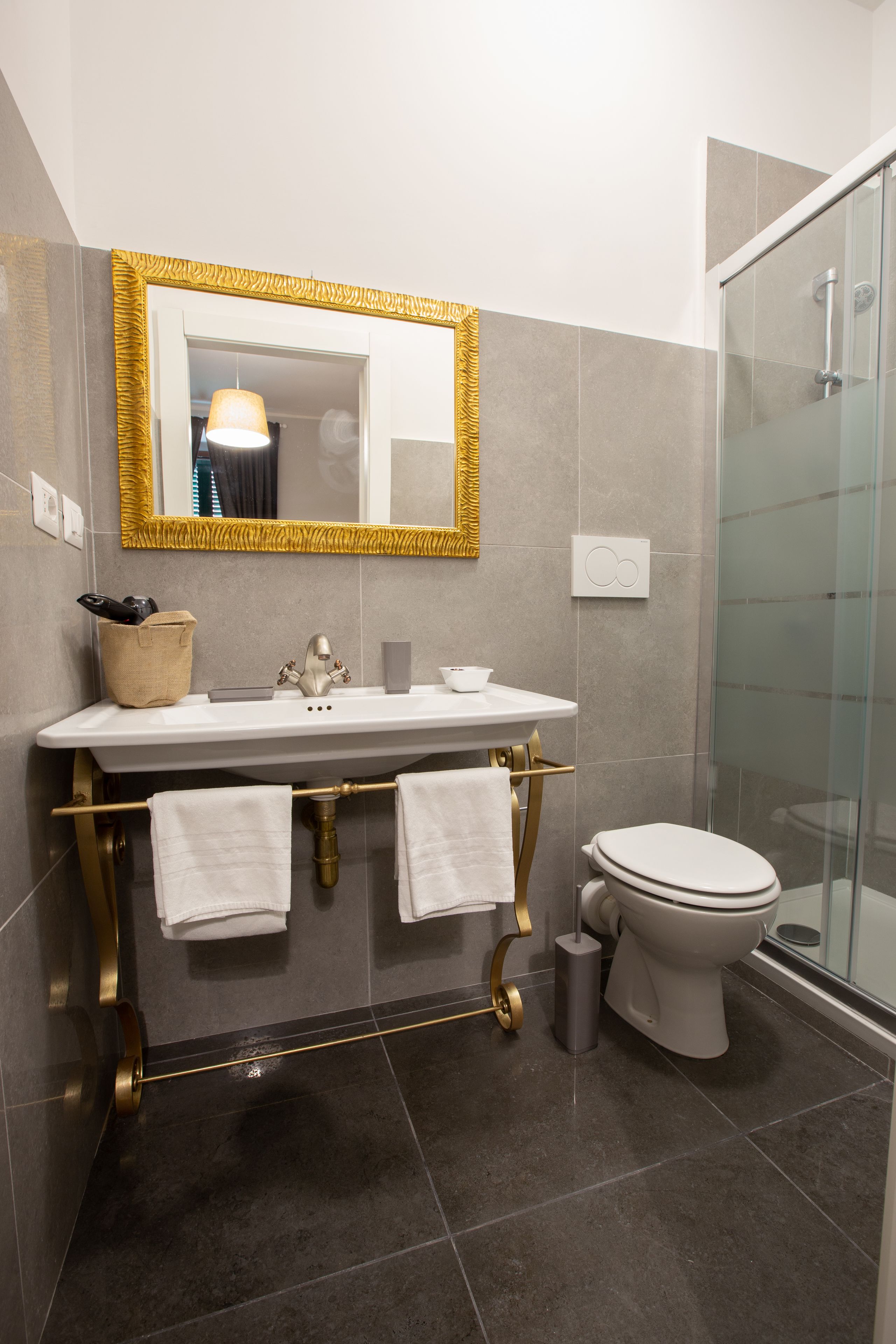 Un baño con lavamanos blanco, grifería dorada, espejo con marco dorado y cabina de ducha de vidrio.