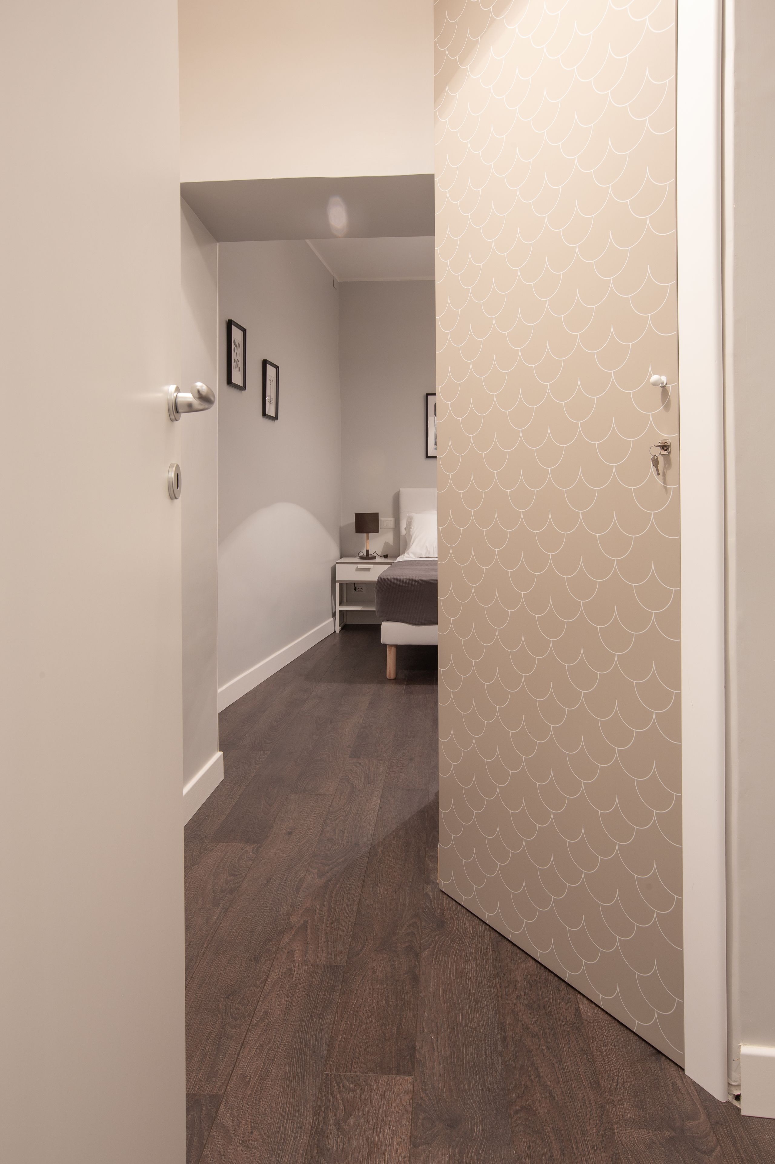 Un couloir avec un plancher en bois, un lit avec une tête de lit blanche et une table de chevet avec une lampe.