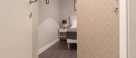 A hallway with a wooden floor, a bed with a white headboard, and a nightstand with a lamp.