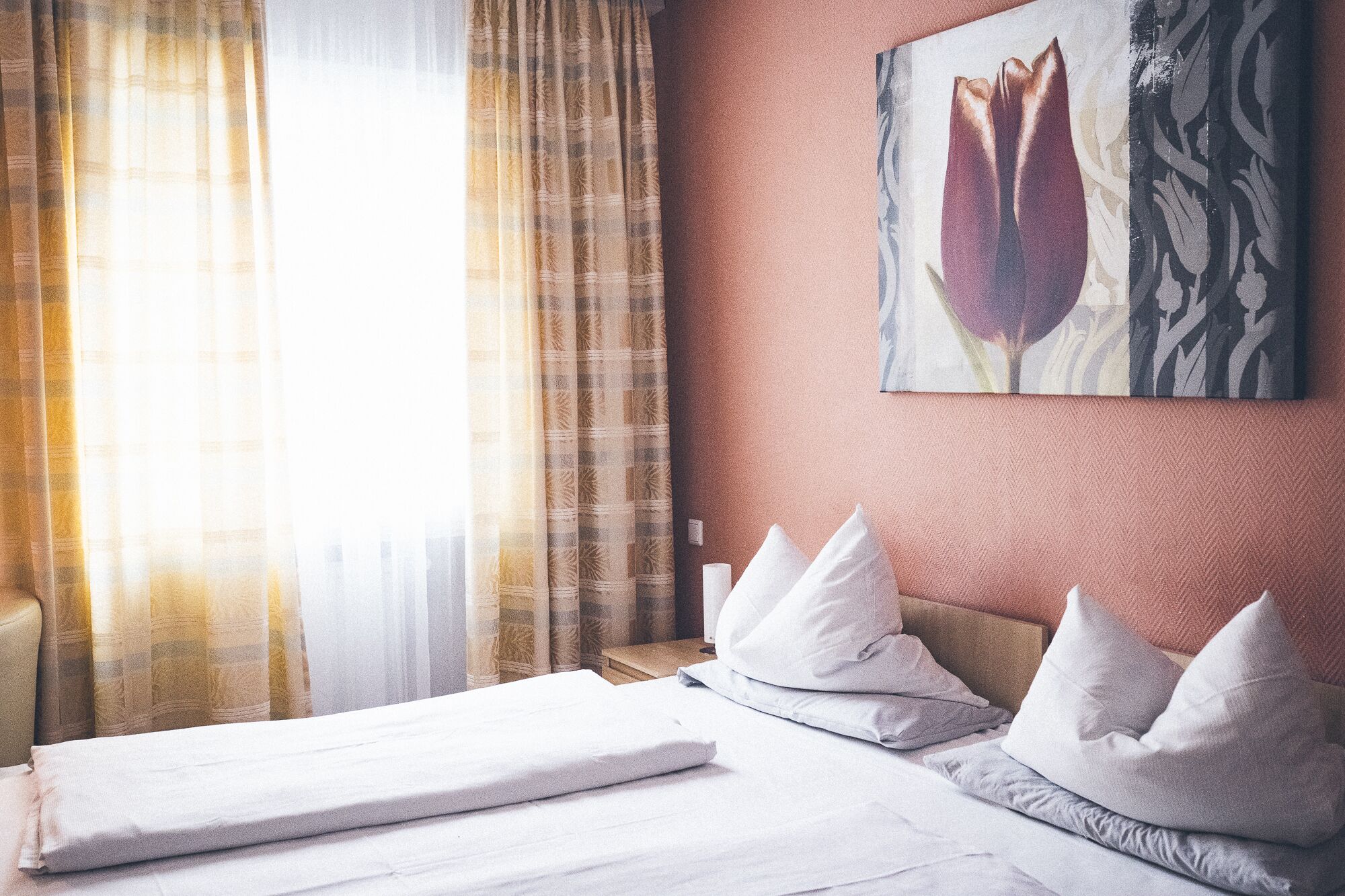Ein Hotelzimmer mit Bett, weißen Kissen, einem Wandbild mit Tulpen und einem Vorhangfenster.