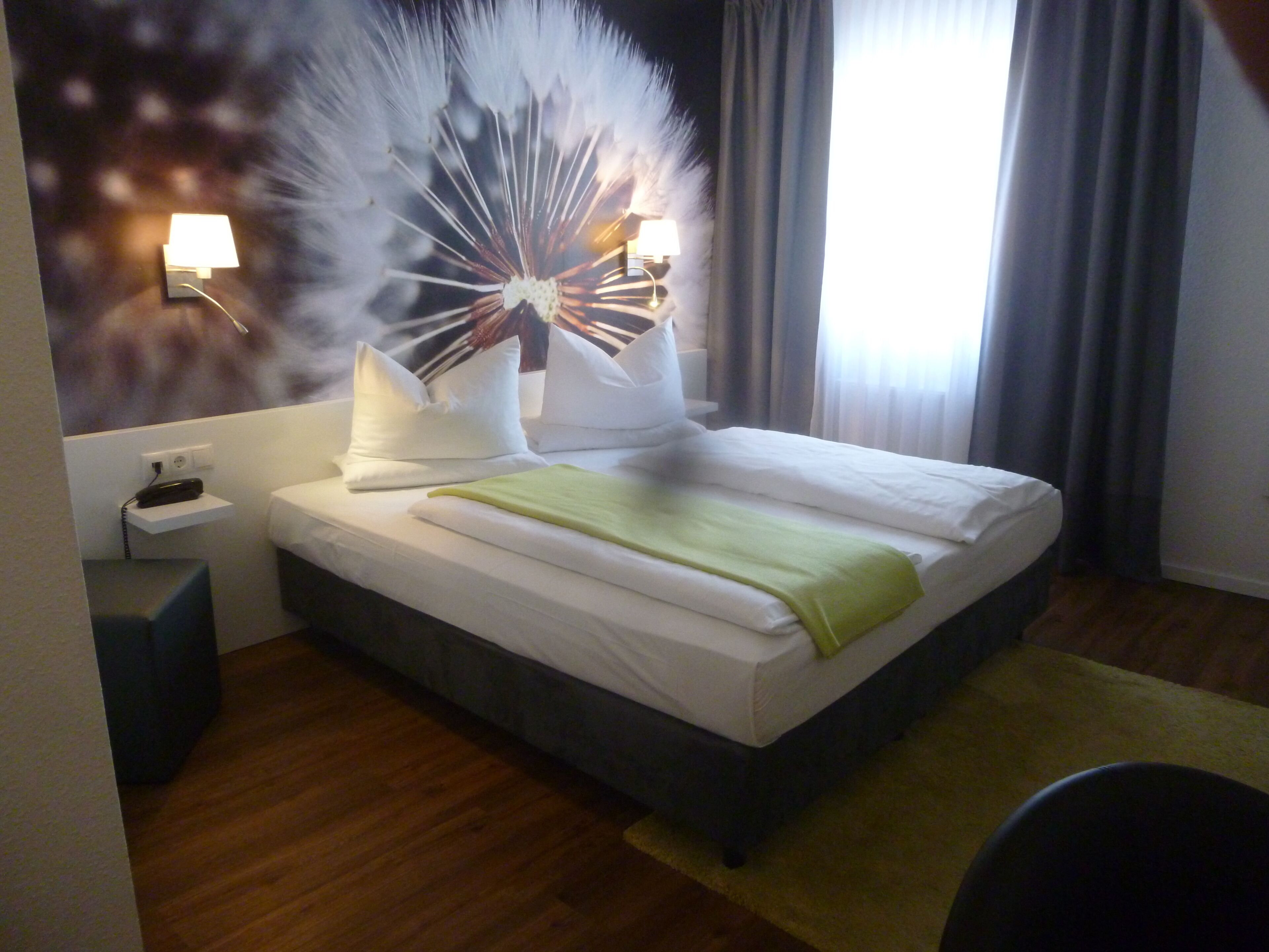Hotellihuone, jossa on sänky, yöpöytä, lamppu ja seinäkoriste.