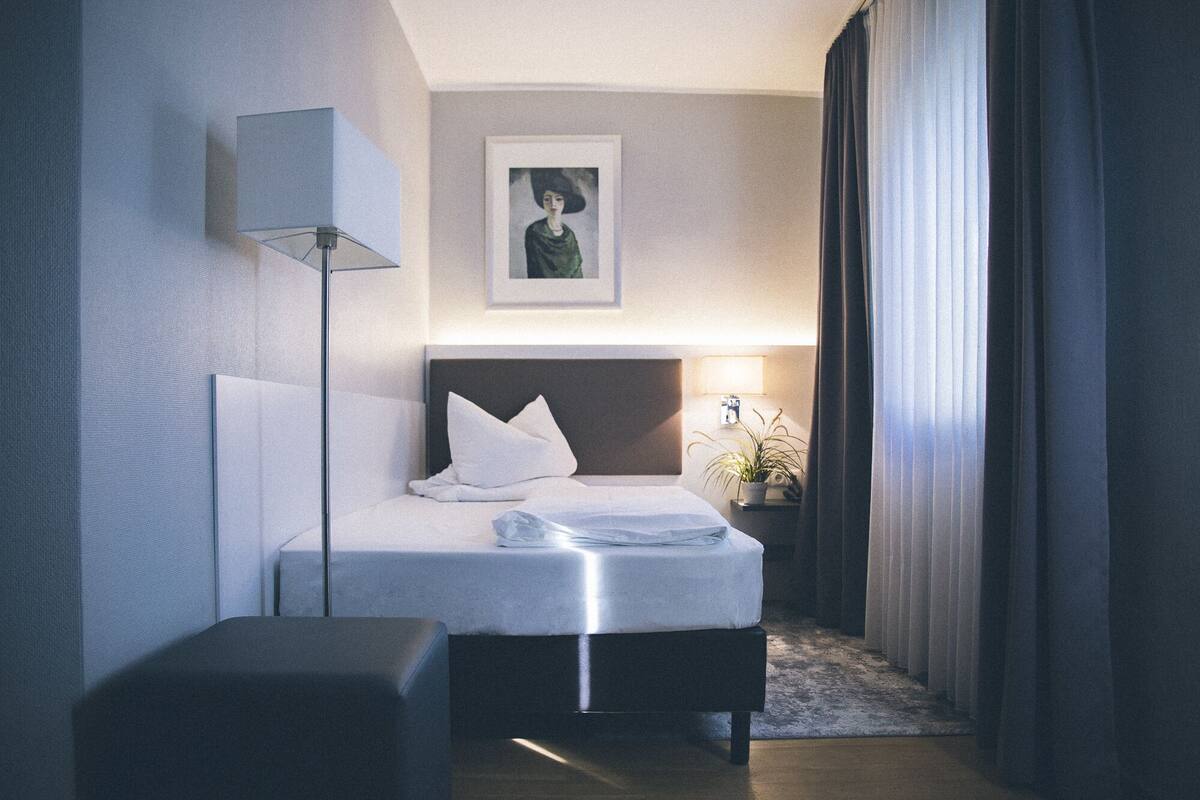 A hotel room with a neatly made bed, a bedside lamp, a framed picture on the wall, and a window with curtains.