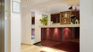 A modern reception area with a wooden reception desk, a red carpet, and a wall-mounted shelf with decorative items. - Gartenstadt Hotel (Ludwigshafen)