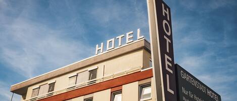 A hotel with a prominent 'HOTEL' sign on the facade and a sign indicating 'Gartenstadt Hotel' with an address.