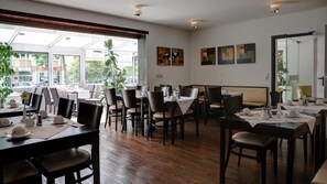 A restaurant with multiple tables and chairs, large windows, and artwork on the walls. - Gartenstadt Hotel (Ludwigshafen)