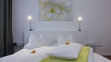Una cama bien hecha con sábanas blancas y un plaid verde, dos mesitas de noche con lámparas, y un cuadro colgado en la pared de flores blancas.