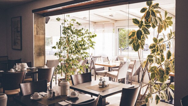 A modern cafe with tables and chairs, large windows, and indoor plants. - Gartenstadt Hotel (Ludwigshafen)