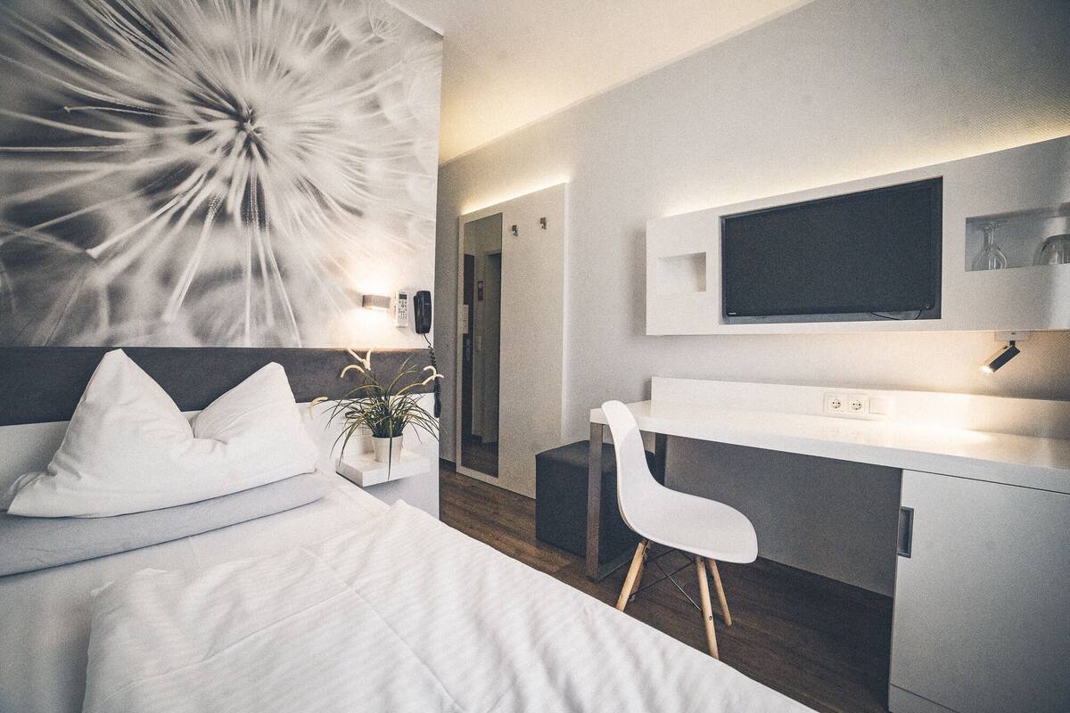 A modern hotel room with a bed, a desk, a chair, a potted plant, and a large abstract artwork on the wall.