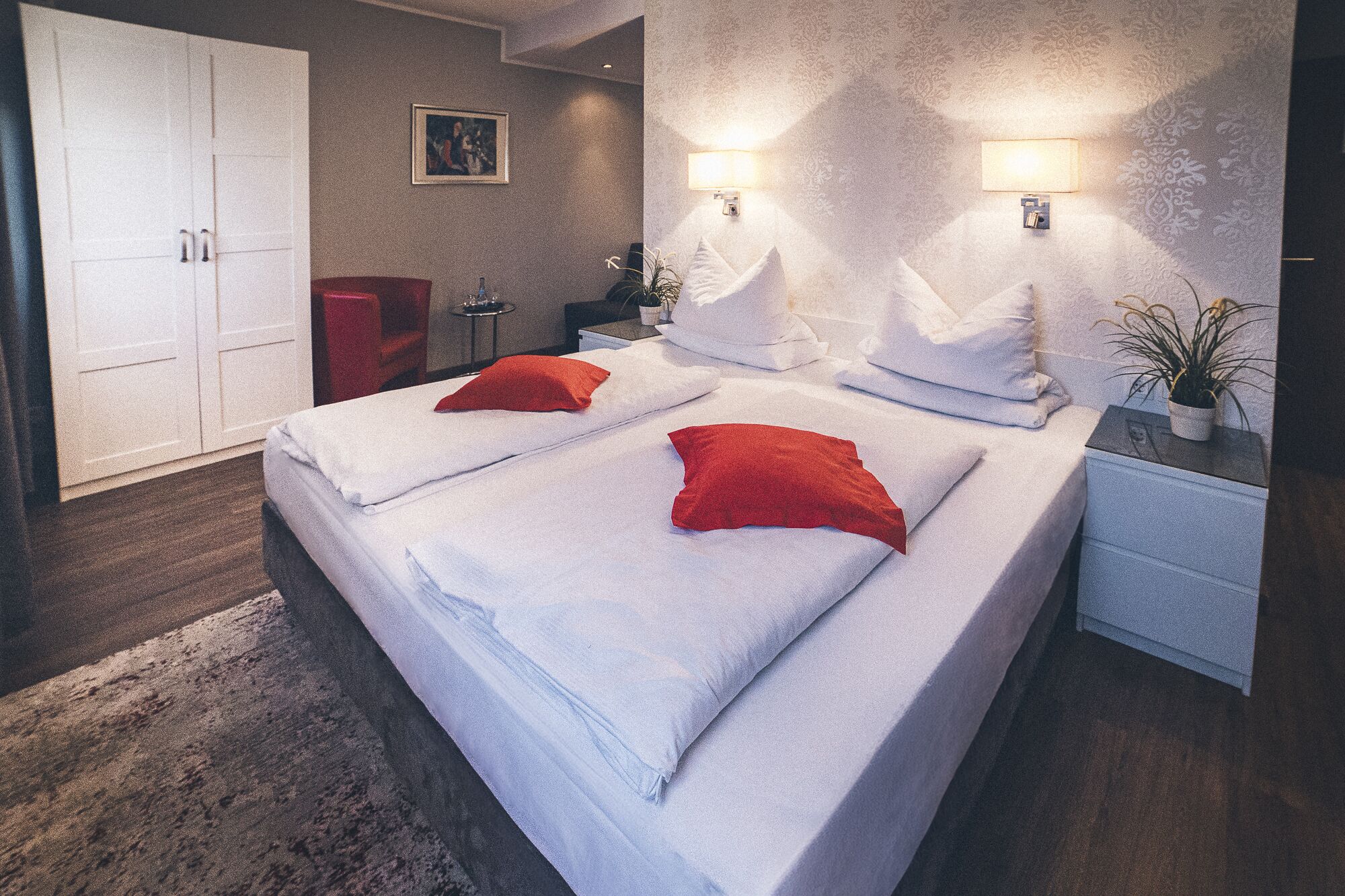 Una cama bien hecha con sábanas blancas y almohadas rojas, una mesita de noche con lámpara, y un armario al fondo.