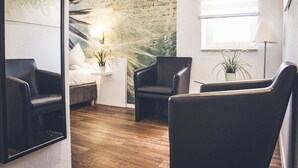 A modern room with a wooden floor, a bed, two black leather chairs, a small table with a plant, and a wall with a nature-themed mural. - Gartenstadt Hotel (Ludwigshafen)