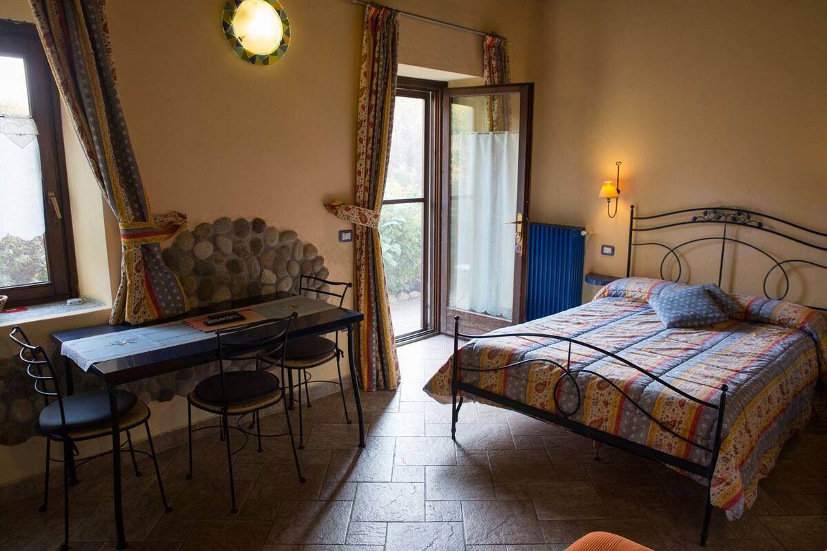 Un dormitorio con cama, una mesa con sillas, una ventana con cortinas y vistas al exterior.