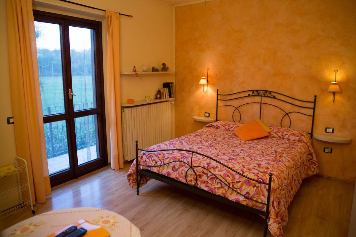 Un dormitorio con una cama, dos lámparas de noche, una ventana con cortinas, un radiador y una estantería con objetos decorativos.