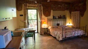 A cozy room with a bed, a table, and a chair. There's a window with curtains, a lamp, and a brick wall. - Agriturismo & Scuderia Fano's Farm (Suno)