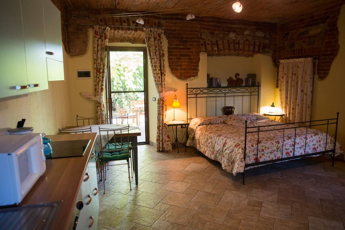 Una habitación acogedora con cama, una mesa y una silla. Se aprecia una ventana con cortinas, una lámpara y una pared de ladrillo.