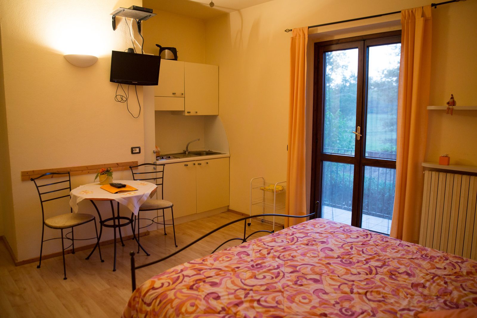 A compact room with a bed, a small dining table, a kitchenette, and a large window.