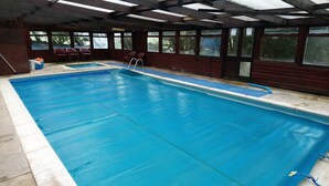 An indoor swimming pool with a clear blue water surface, wooden deck, and large windows offering views of the surrounding area.