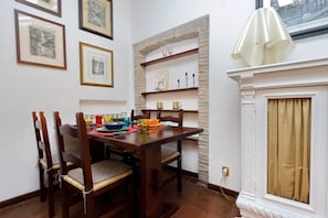A dining area with a wooden table and chairs, framed artwork on the walls, a shelf with decorative items, and a fireplace with a curtain.