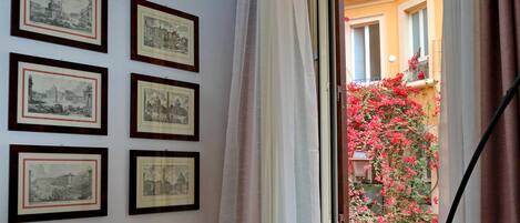 A room with six framed black and white photographs on the wall, a window with a view of a building with balconies and red flowers, and a curtain partially drawn.