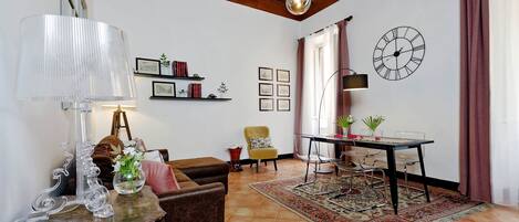 Ein Wohnzimmer mit Holzboden, einem Essbereich mit Tisch und Stühlen, einer Wanduhr und einem Sofa mit Lampe.