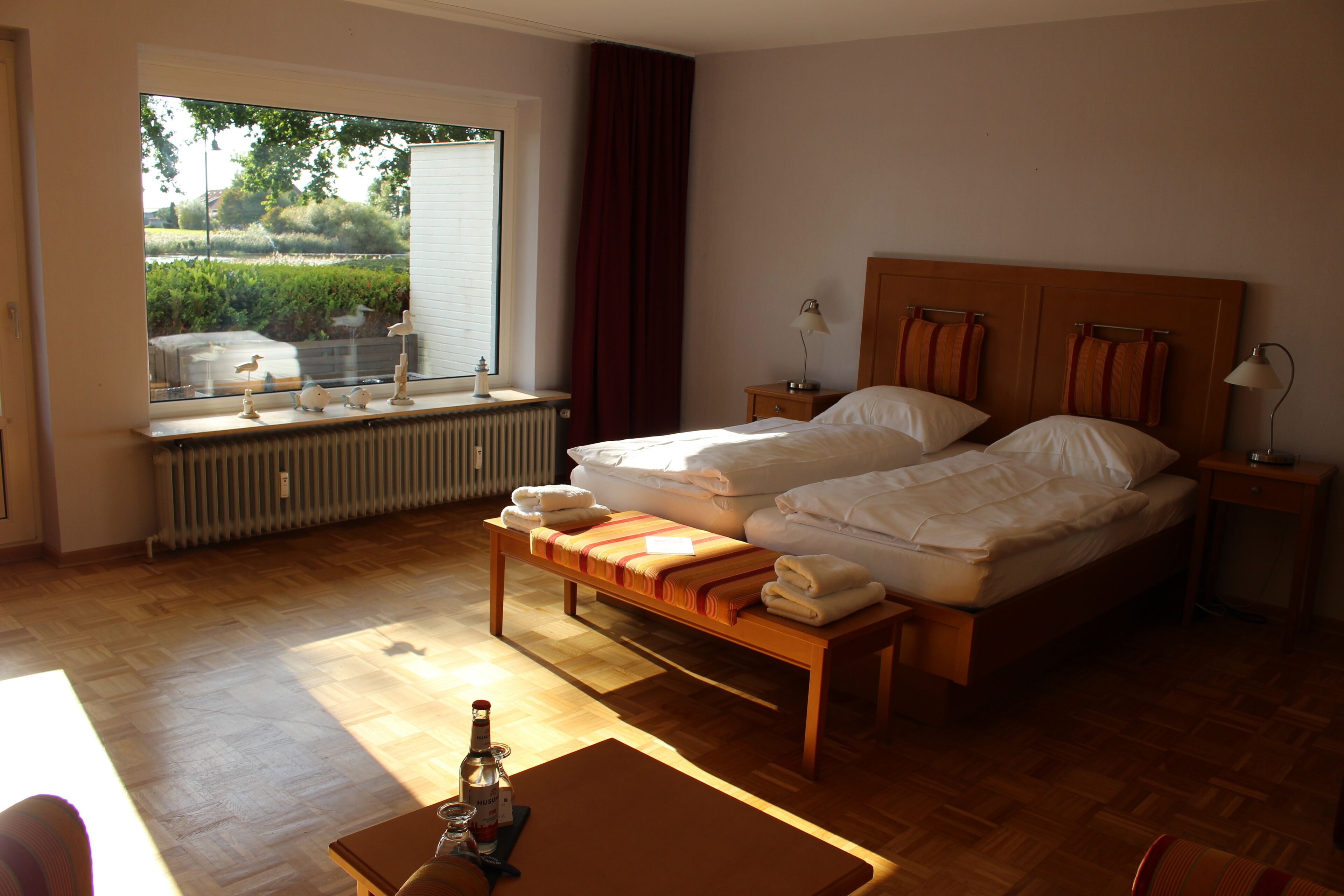 Habitación de hotel con una cama grande, un banco, una mesita con una botella y una ventana con vistas a zonas verdes.