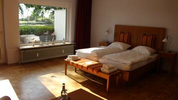 Habitación de hotel con una cama grande, un banco, una mesita con una botella y una ventana con vistas a zonas verdes.