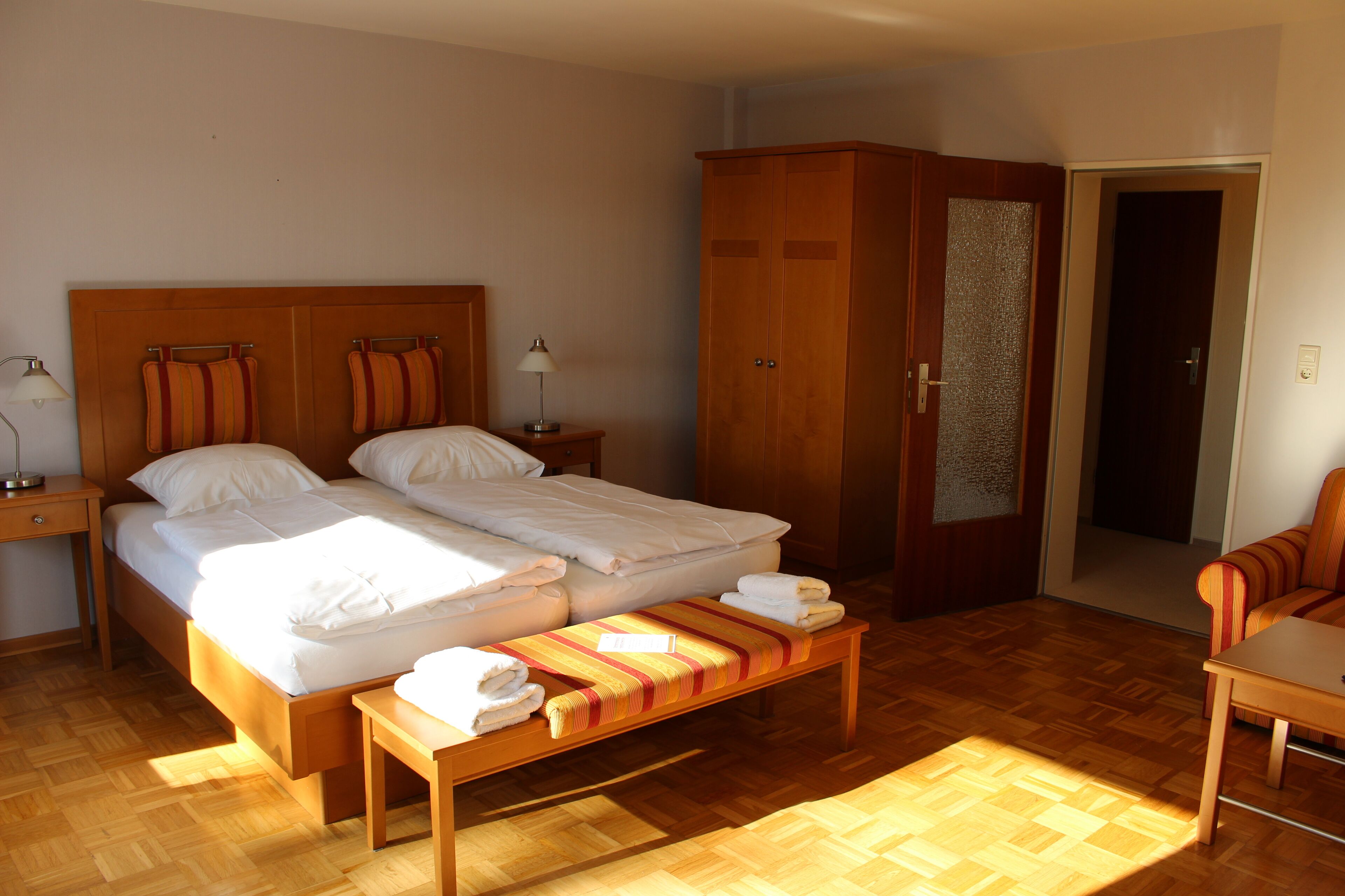 A bedroom with a double bed, a wooden wardrobe, a bench with towels, and a small table.