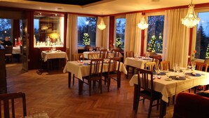 A dining area with multiple tables set for a meal, large windows with curtains, and chandeliers. - Hotel Zur Treene (Schwabstedt)