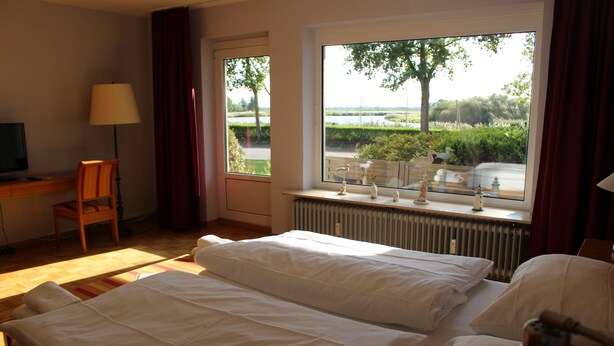 A hotel room with two beds, a desk, a chair, a TV, and a window with a view of greenery and a body of water. - Hotel Zur Treene (Schwabstedt)