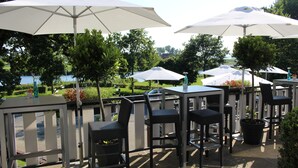 An outdoor patio area with tables and chairs under large umbrellas, surrounded by greenery and a view of a lake. - Hotel Zur Treene (Schwabstedt)