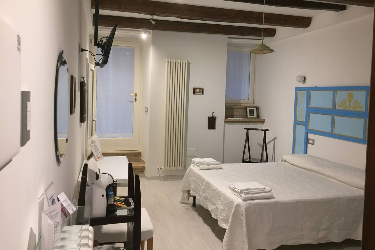 A room with a bed, a desk with a chair, a radiator, a window, and a blue-framed door.