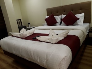 A neatly made bed with white and red bedding, a brown headboard, and a bedside table with a lamp and a water bottle. - Hotel Alfa International (Mumbai)
