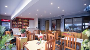 A restaurant with wooden tables and chairs, a Coca-Cola refrigerator, and shelves with bottles and glasses. - Hotel Rosemary Homes (Kathmandu)