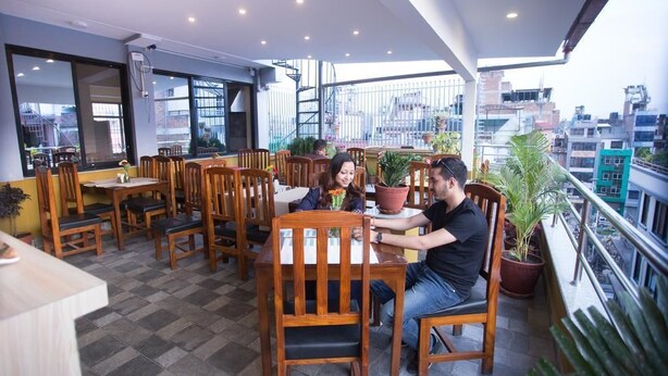An indoor dining area with wooden tables and chairs, large windows, and potted plants. - Hotel Rosemary Homes (Kathmandu)