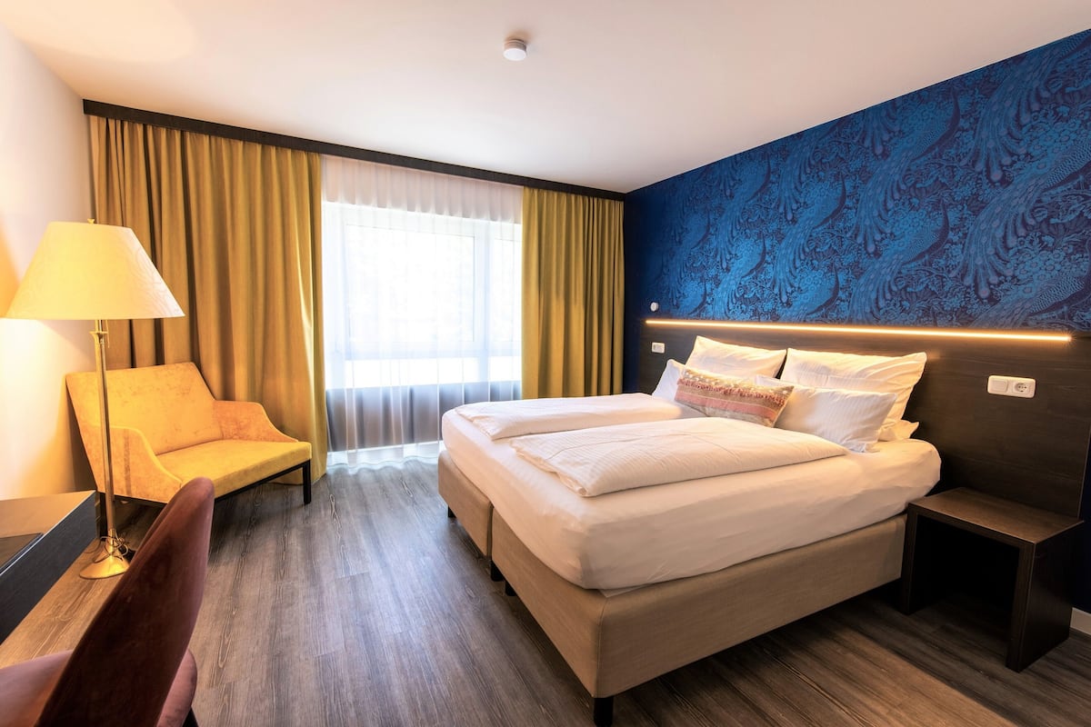 Habitación de hotel con cama doble, escritorio con lámpara, silla y ventana grande con cortinas.