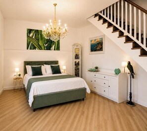 A bedroom with a bed, bedside tables, a dresser, a chandelier, a staircase, and a wall with a plant and a painting.