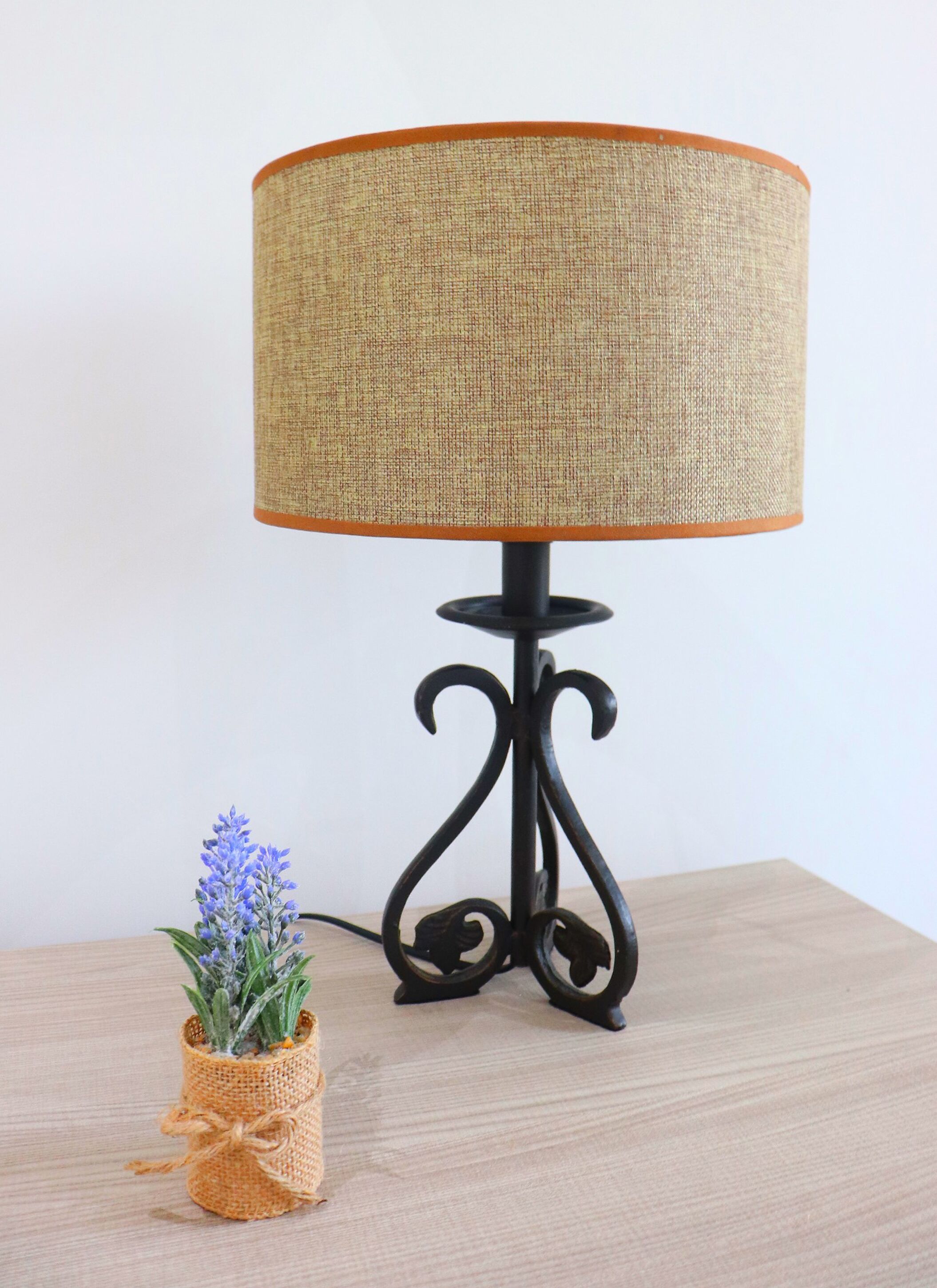 A table lamp with a woven shade and a black metal base, placed on a wooden table next to a small plant in a woven pot.