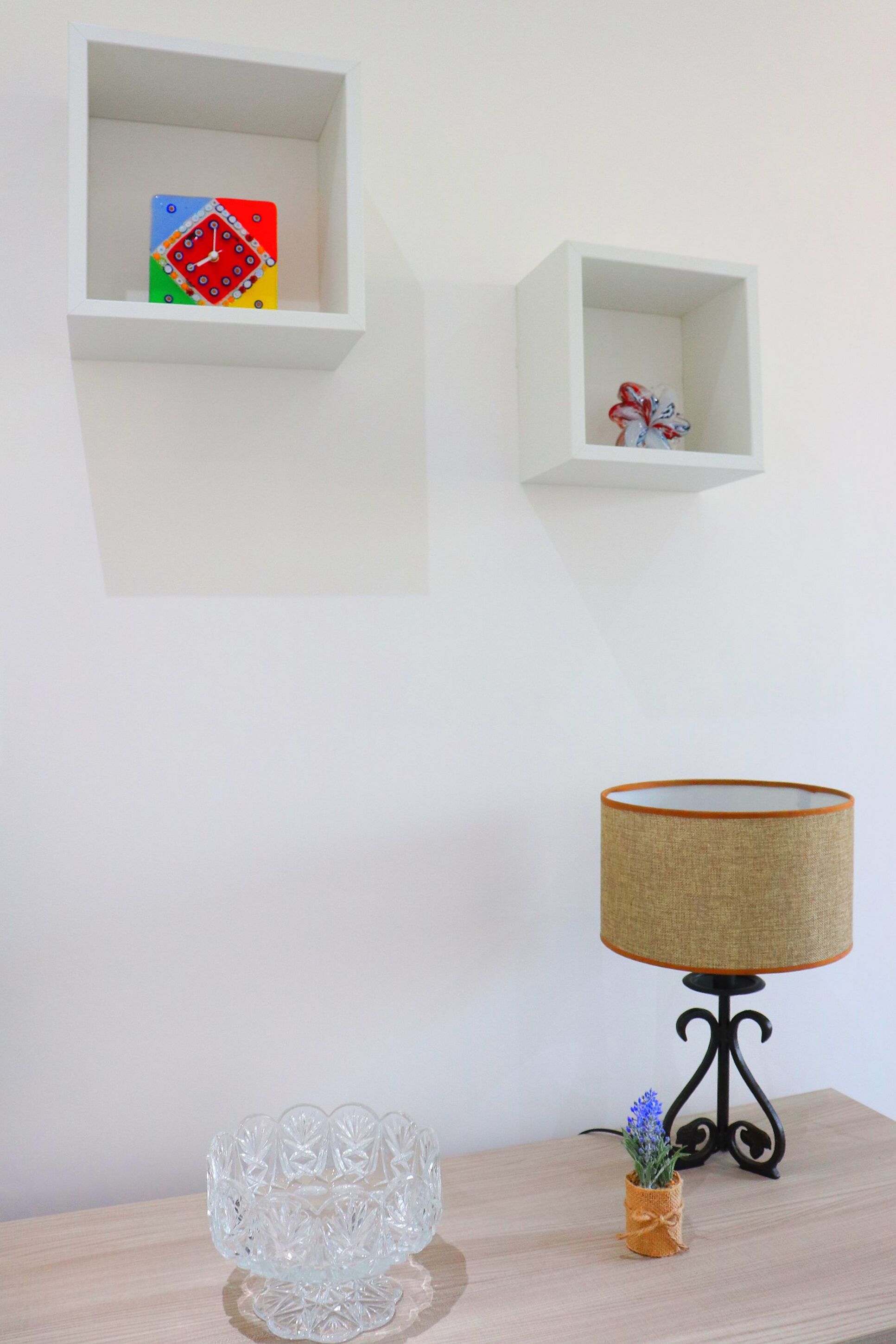 A room with a wooden table, a clear glass vase, a lamp, and two white shelves with decorative items.