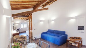 A modern living room with a blue sofa, wooden ceiling beams, and a dining area with a glass table. - Domizio - WR Apartments (Rome)