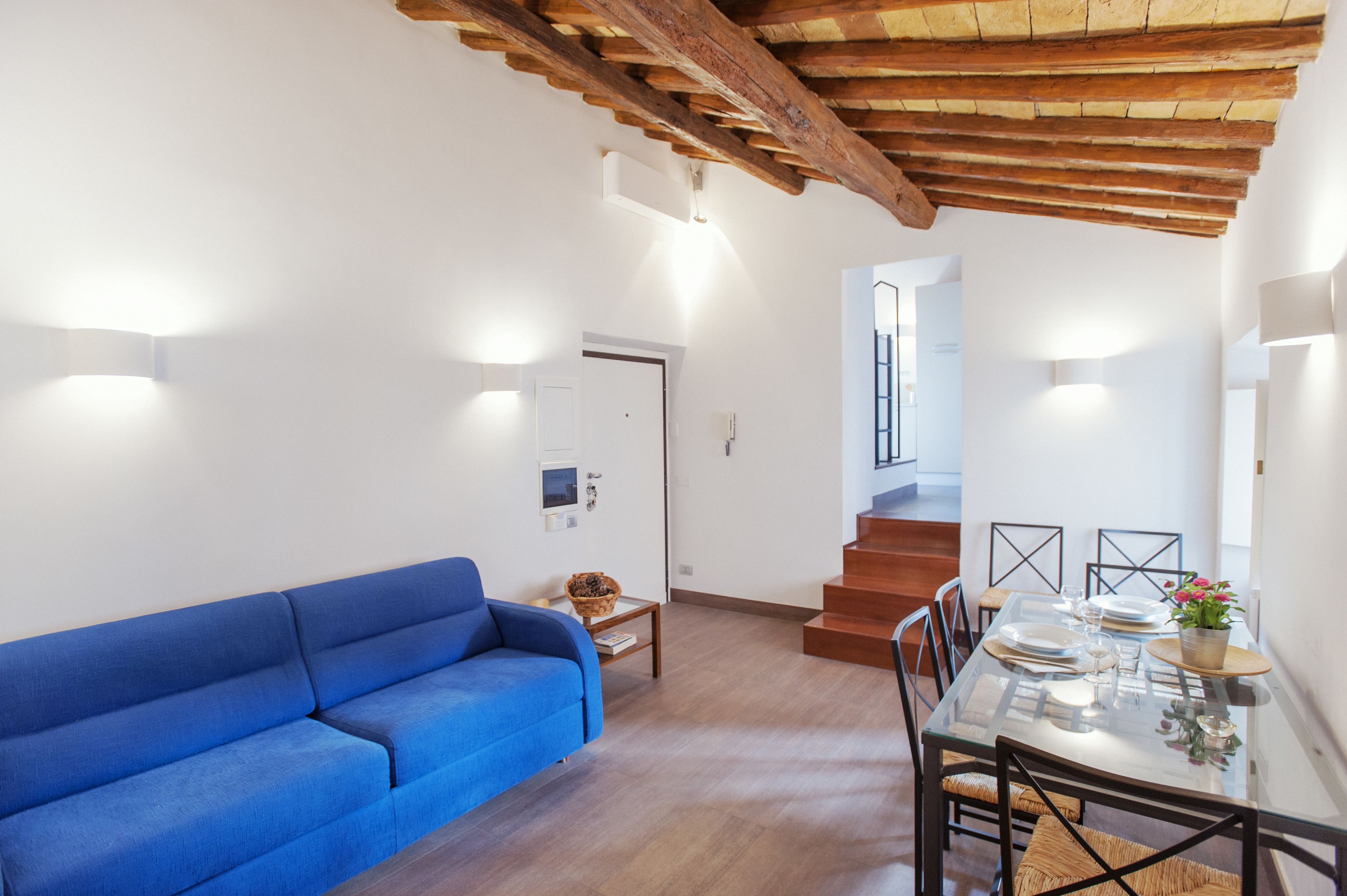 Una sala de estar moderna con un sofá azul, una mesa de comedor de vidrio preparada para cuatro personas y una escalera de madera que sube a un nivel superior.
