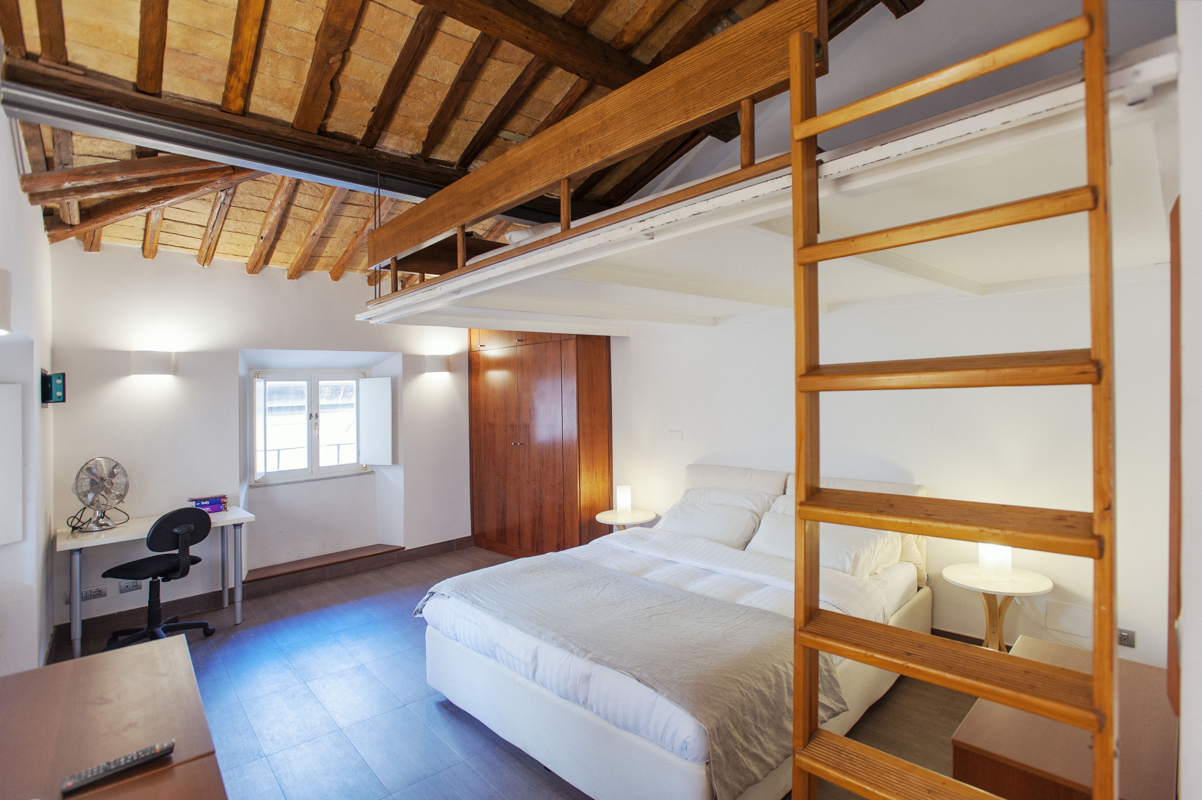 A bedroom with a bunk bed, a desk with a chair, a small table, and a fan.