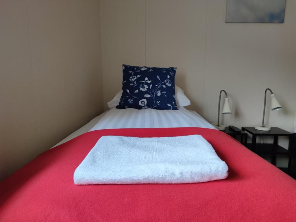 A neatly made bed with a red bedspread and a white towel, a blue floral pillow, and two bedside lamps on a nightstand.
