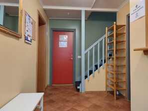 Un couloir avec une porte rouge, un escalier en bois et un panneau d’affichage contenant des informations sur les différentes pièces.