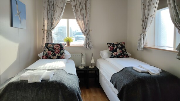 Une chambre avec deux lits simples, chacun agrémenté d’un coussin à motifs floraux et d’une couverture foncée. On y trouve une petite table avec une lampe placée entre les lits, ainsi qu’une fenêtre ornée de rideaux à motifs floraux.