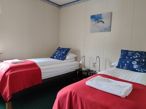 Une chambre d’hôtel avec deux lits, chacun recouvert d’une couverture rouge et agrémenté d’un coussin à motifs floraux bleus. Une table de chevet avec une lampe se trouve entre les deux lits. Un tableau représentant un oiseau est accroché au mur, au-dessus des lits.