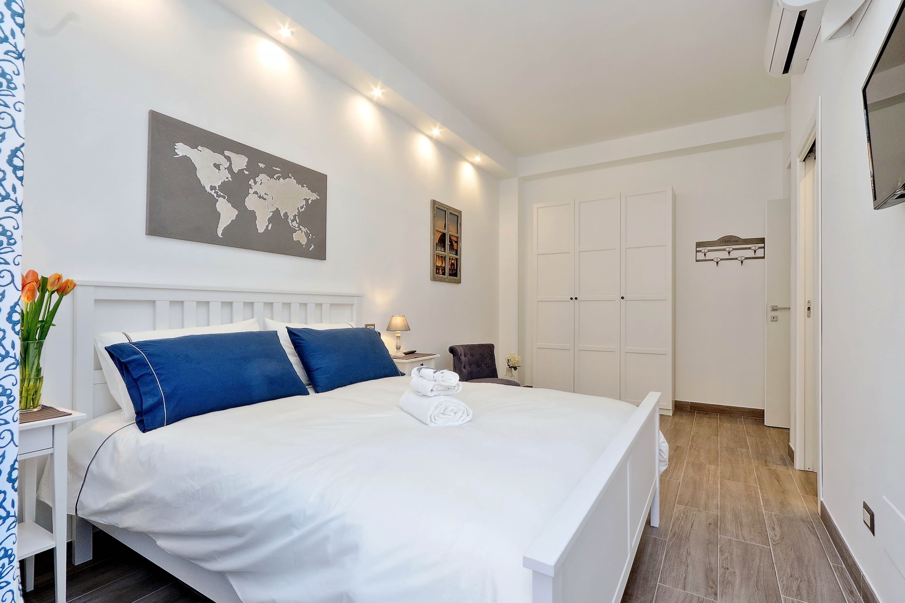 Una cama bien tendida con ropa de cama blanca y azul, un mapa mundial en la pared y una mesita con una lámpara y flores.