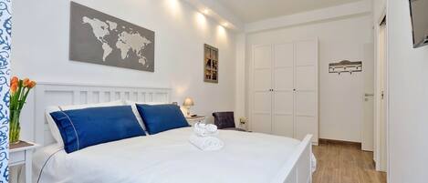 A neatly made bed with white and blue bedding, a world map on the wall, and a small table with a lamp and flowers.